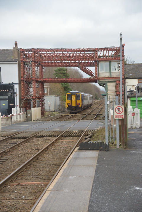 A rural scene
Class 156 trainset 156414 draws away from Bamber Bridge whilst working a Preston - Colne local service. 
NOTE the ironwork supporting the crossing box (once used a the basis for a model signal box on model railways) now providing a frame for the image.
